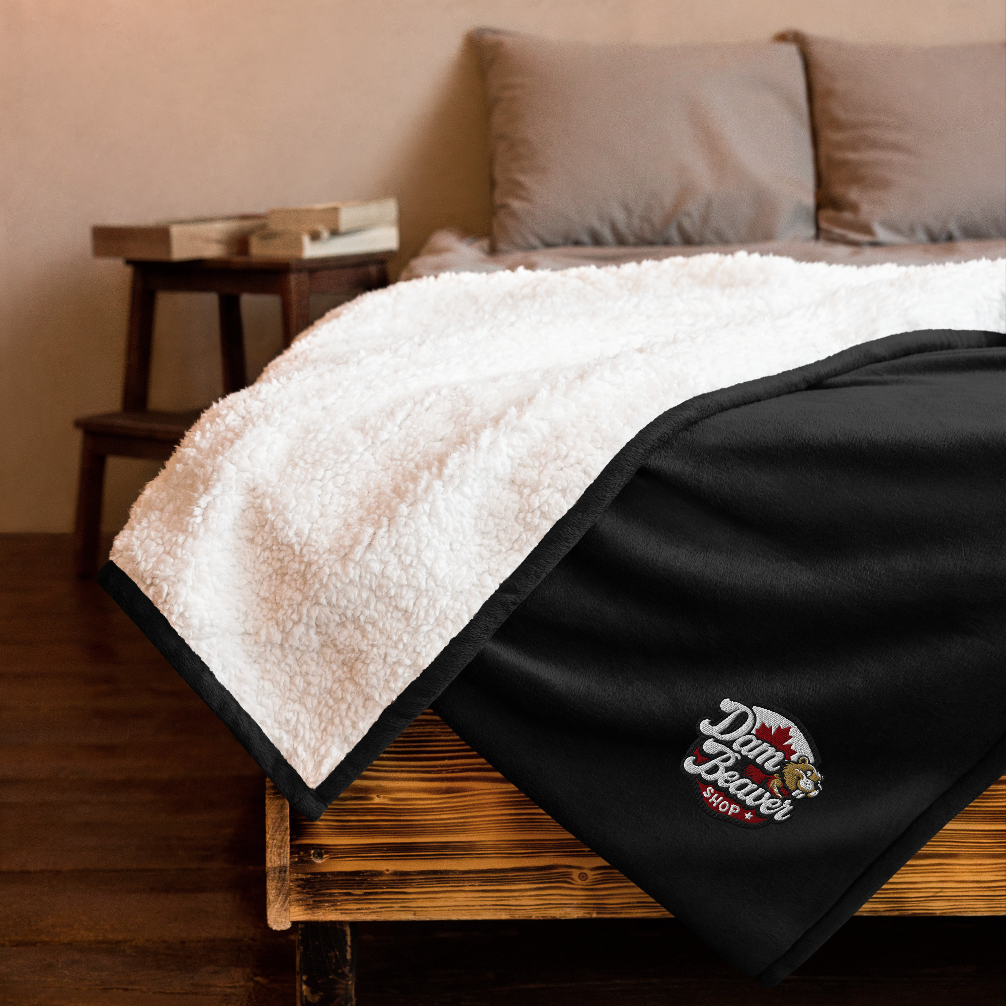 A cozy bed features the Cozy Embroidered Premium Sherpa Blanket - 50 x 60 Comfort in white and black, with the Dam Beaver Shop logo embroidered on one corner. Pillows and a wooden nightstand are visible in the background.